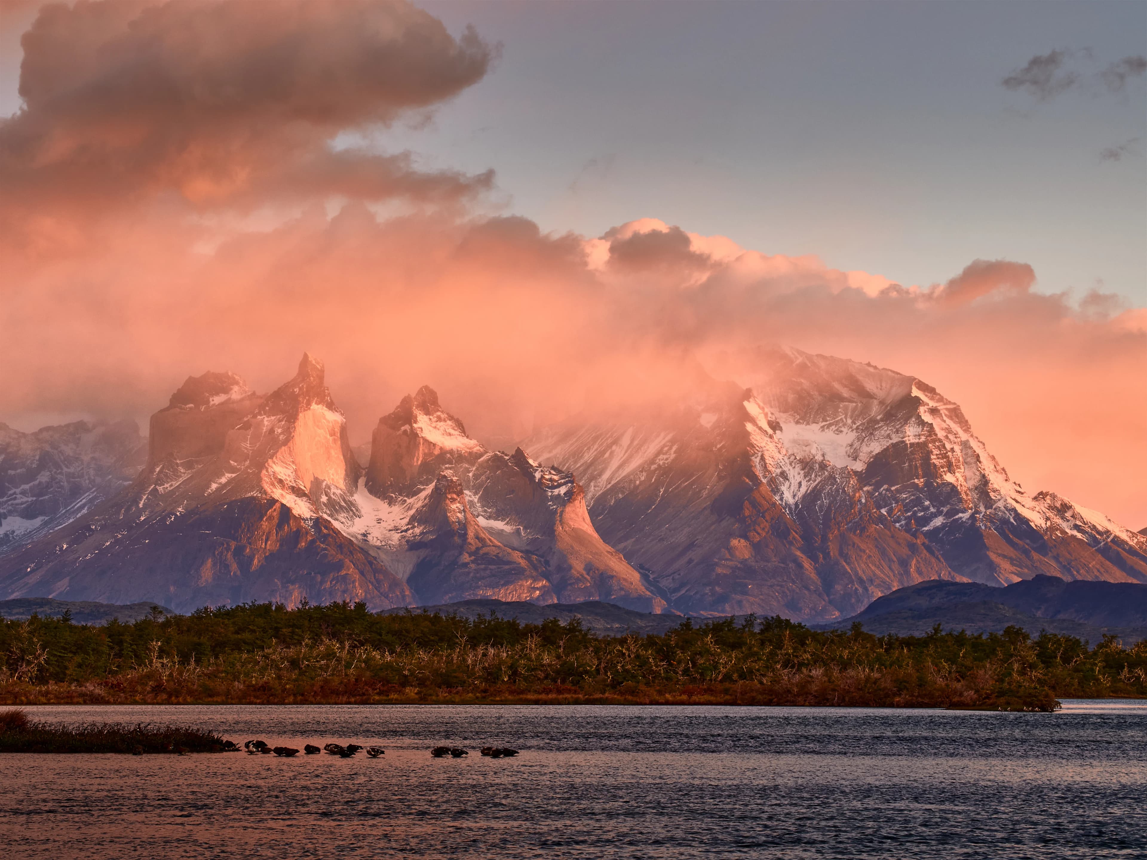 The jagged Cuernos del Paine rise above a blue lake and tawny grass beneath broken cloud.