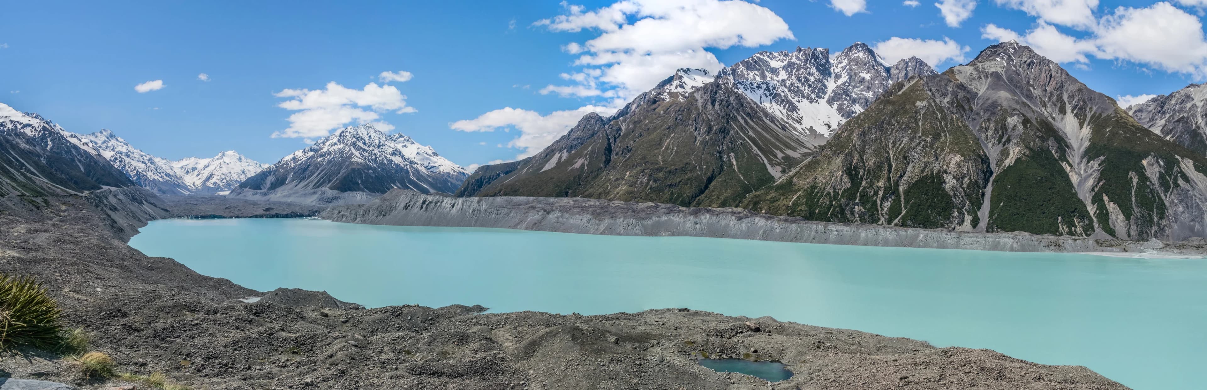 A wide panorama of pale blue Tasman Lake, glacial moraine and snow-covered mountains beneath clear sky.