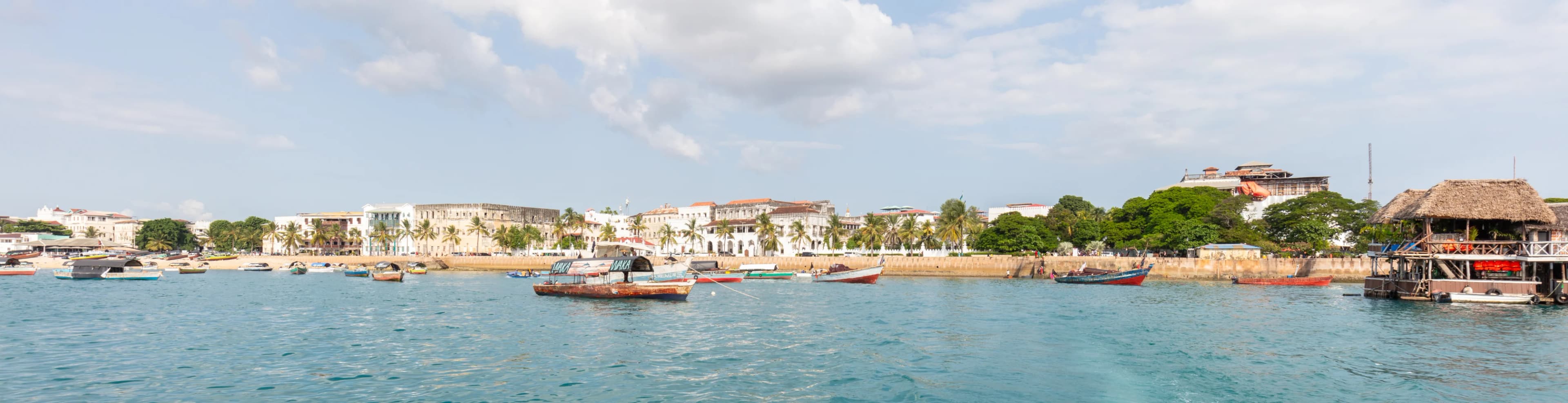 A panoramic view of Stone Town waterfront in Zanzibar, with boats crossing calm blue water.